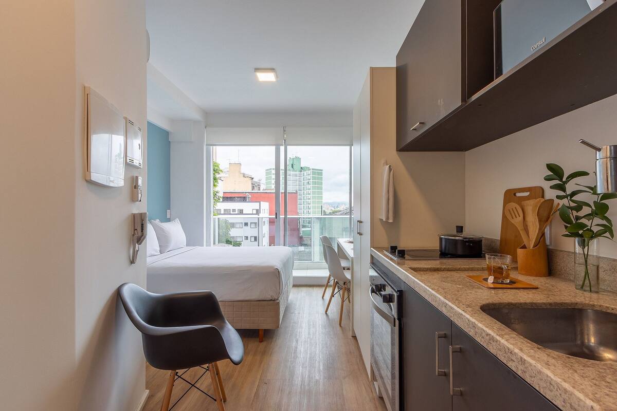 Une chambre d’hôtel compacte comprenant un lit, un petit bureau, une kitchenette et une grande fenêtre donnant sur la ville.