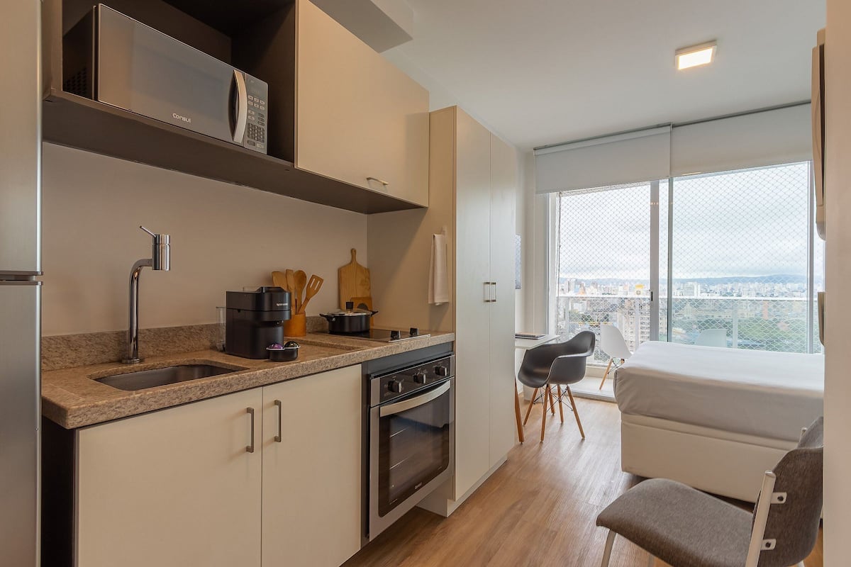 Une cuisine moderne équipée d’un four encastré, d’un four à micro-ondes et d’une machine à café. Une fenêtre offre une vue sur la ville, et un petit coin salon avec une chaise et une table est également disponible.