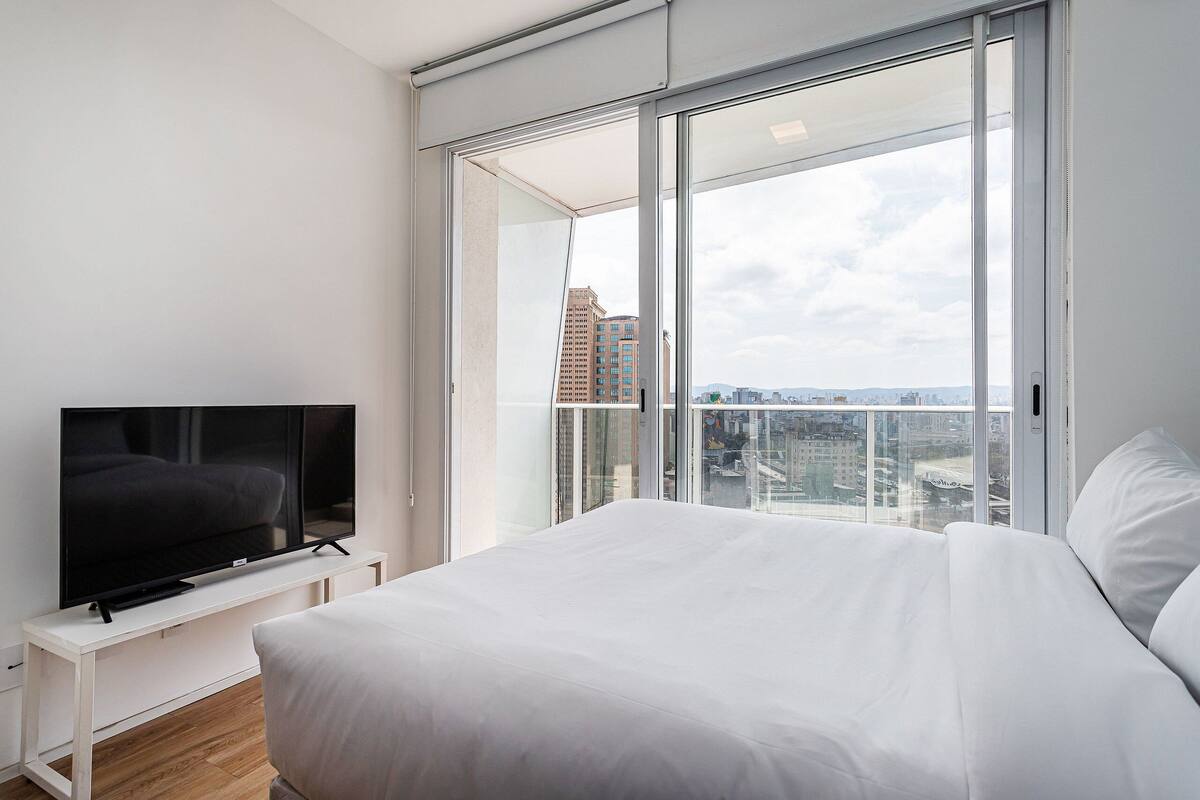 Une chambre moderne avec un grand lit, un téléviseur posé sur une console blanche et une vue sur la ville grâce à de grandes fenêtres du sol au plafond.