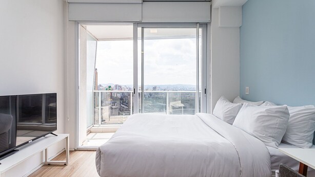 Un dormitorio moderno con una cama grande, un televisor sobre un mueble blanco y un balcón con vista a la ciudad.