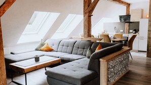 Un espace de vie de style loft comprenant un canapé d’angle, une table basse et un banc intégré. On y trouve également des fenêtres de toit, un coin cuisine avec une cuisinière et un évier, ainsi qu’un téléviseur fixé au mur.