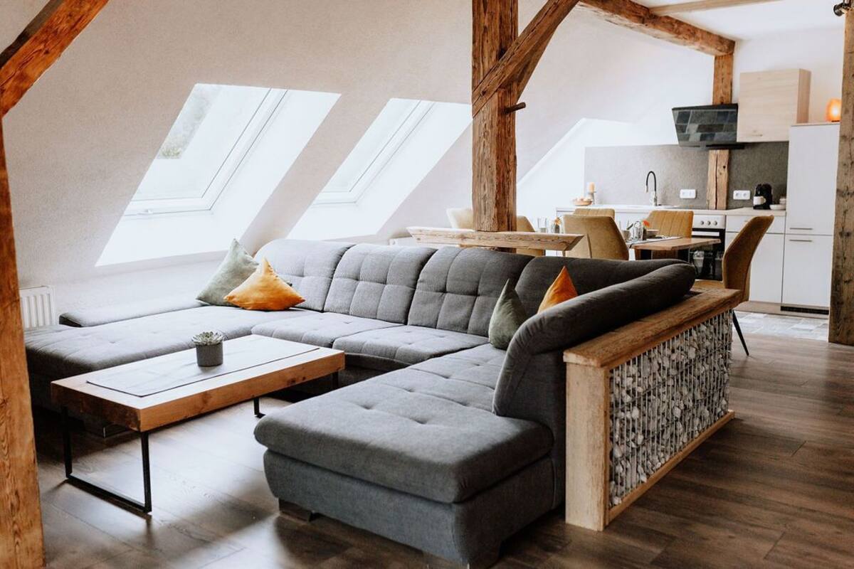 Un espace de vie de style loft comprenant un canapé d’angle, une table basse et un banc intégré. On y trouve également des fenêtres de toit, un coin cuisine avec une cuisinière et un évier, ainsi qu’un téléviseur fixé au mur.