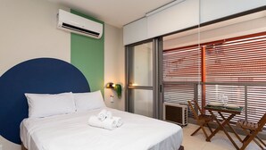 Hotelkamer met een bed, een groot blauw hoofdbord, een airconditioner, een balkon met houten zonwering en een tafel met stoelen.