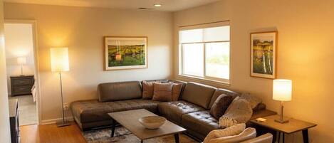 A modern living room with a sectional sofa, a coffee table, and two side tables with lamps. There are framed pictures on the walls and a large window providing natural light.