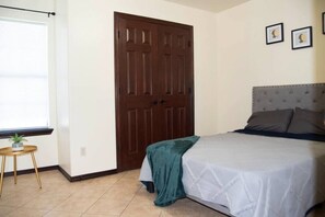 Une chambre à coucher avec un lit, une porte en bois, une petite table avec une plante et une fenêtre avec des stores.