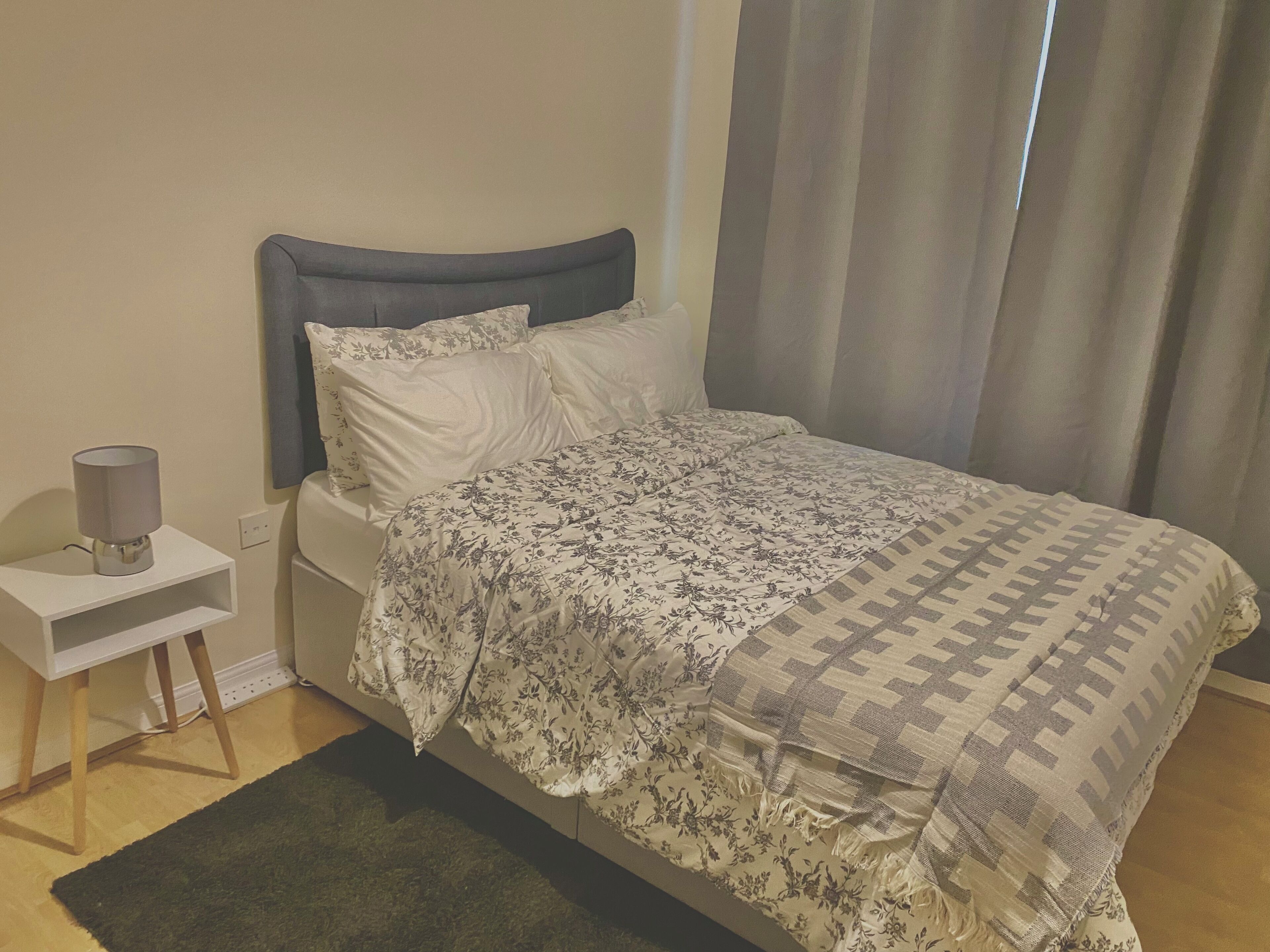 Un dormitorio con cama, mesita de noche, lámpara y ventana con cortinas.