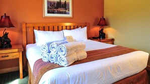 Una cama bien hecha con toallas enrolladas y una manta, dos mesitas de noche con lámparas, y un cuadro enmarcado en la pared.