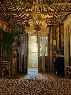 Une pièce avec un plafond en bambou, un lustre suspendu en macramé, une grande plante en pot et un tapis tissé.