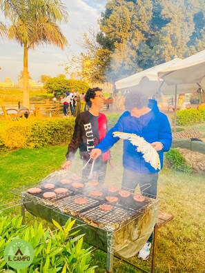 Una parrilla de barbacoa con hamburguesas cocinándose al aire libre, rodeada de follaje y una carpa.