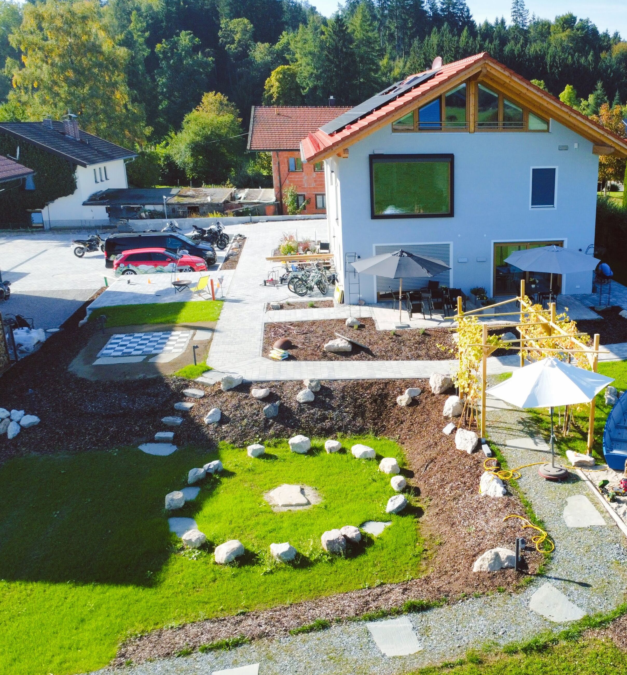 Una casa moderna con jardín, un patio con sombrillas y un área de estacionamiento con coches.