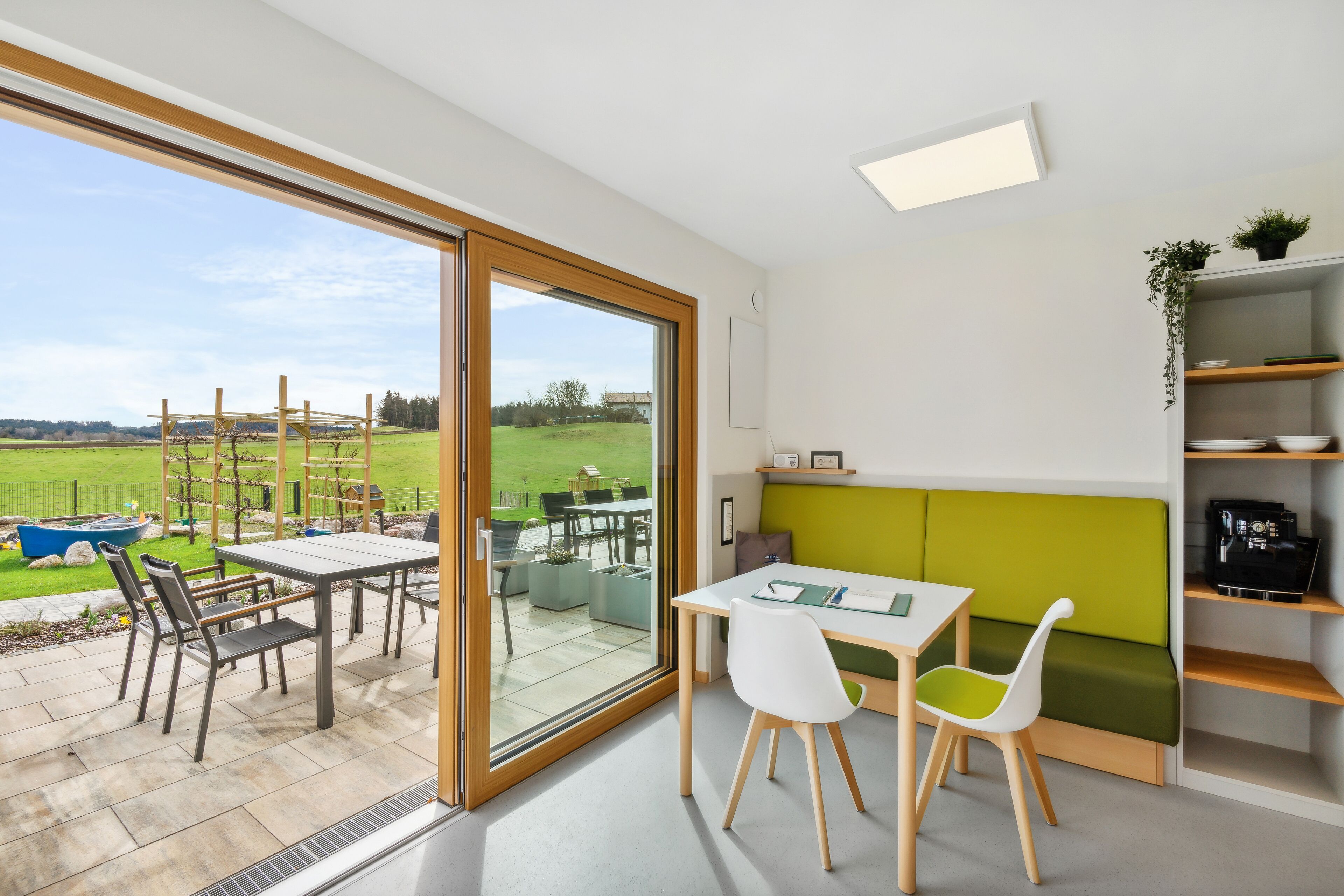 Cocina moderna con una decoración verde y blanca, una zona de comedor con mesa y sillas, y un gran ventanal corredero que da a un patio con mobiliario exterior.
