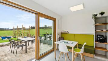 Cocina moderna con una decoración verde y blanca, una zona de comedor con mesa y sillas, y un gran ventanal corredero que da a un patio con mobiliario exterior.