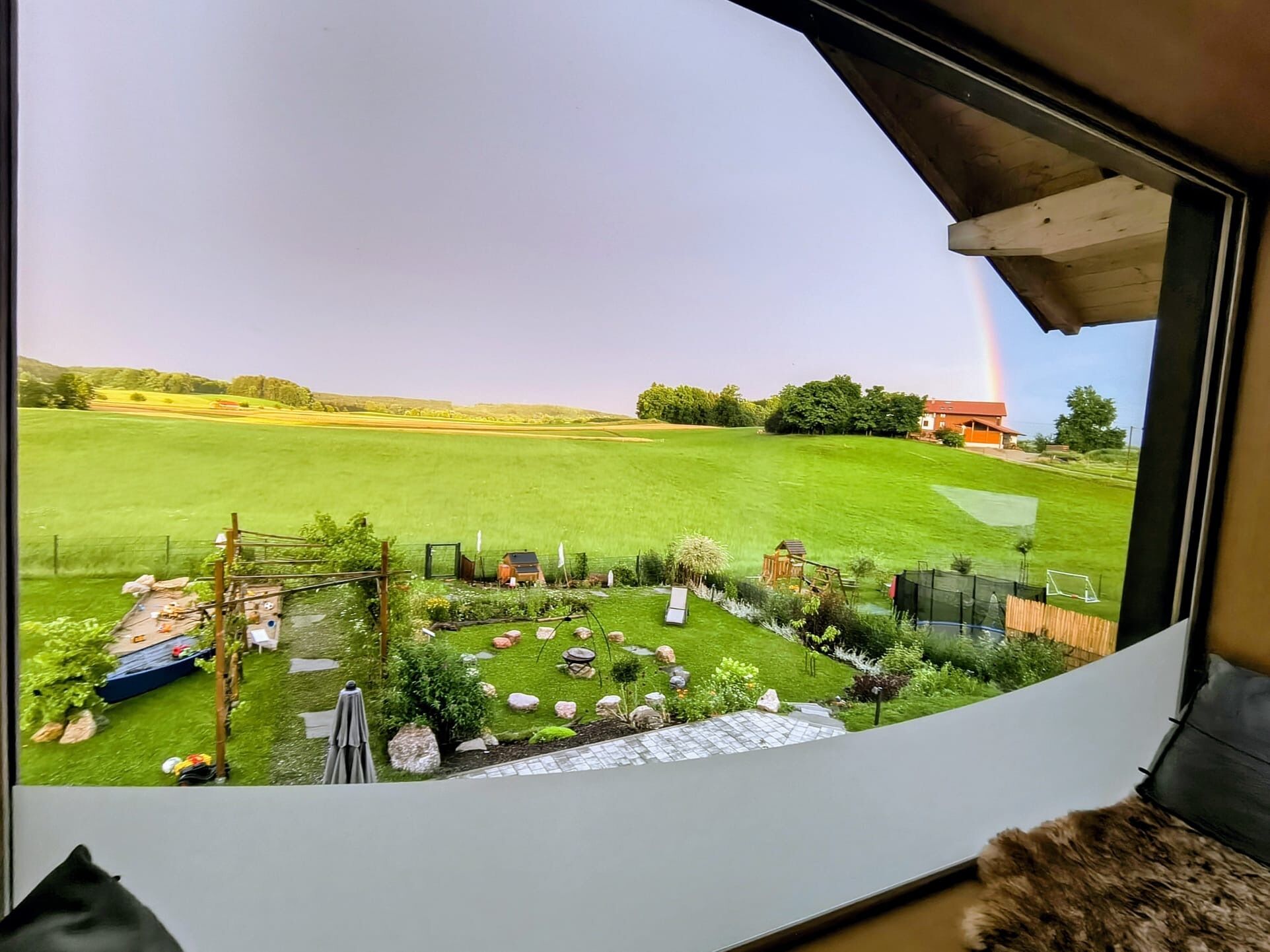 A view from a window of a room with a view of a grassy field, a small wooden structure, and a rainbow in the distance.