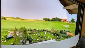 A view from a window of a room with a view of a grassy field, a small wooden structure, and a rainbow in the distance.