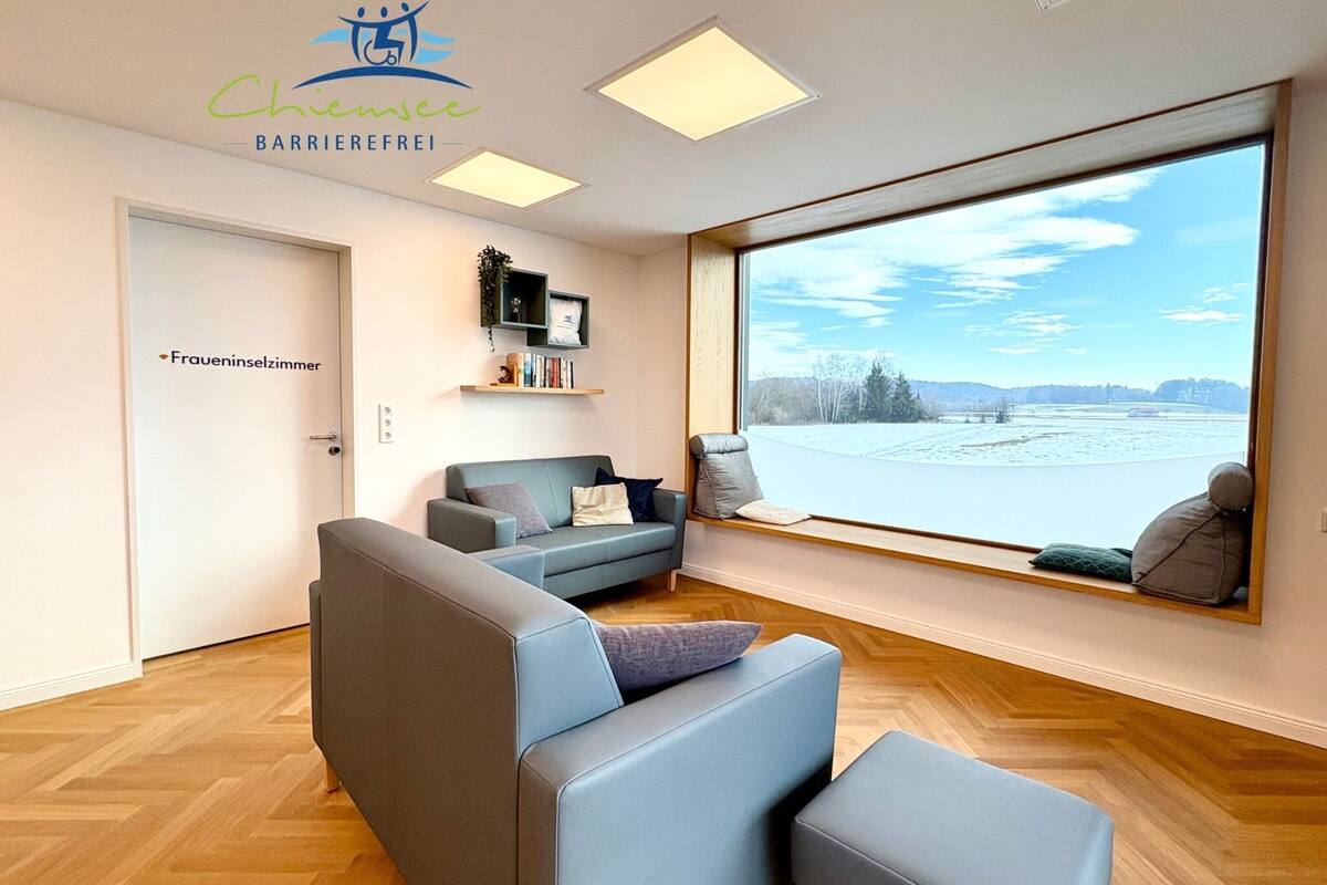 A modern living room with a sofa, ottoman, and a large window offering a view of a river and trees.