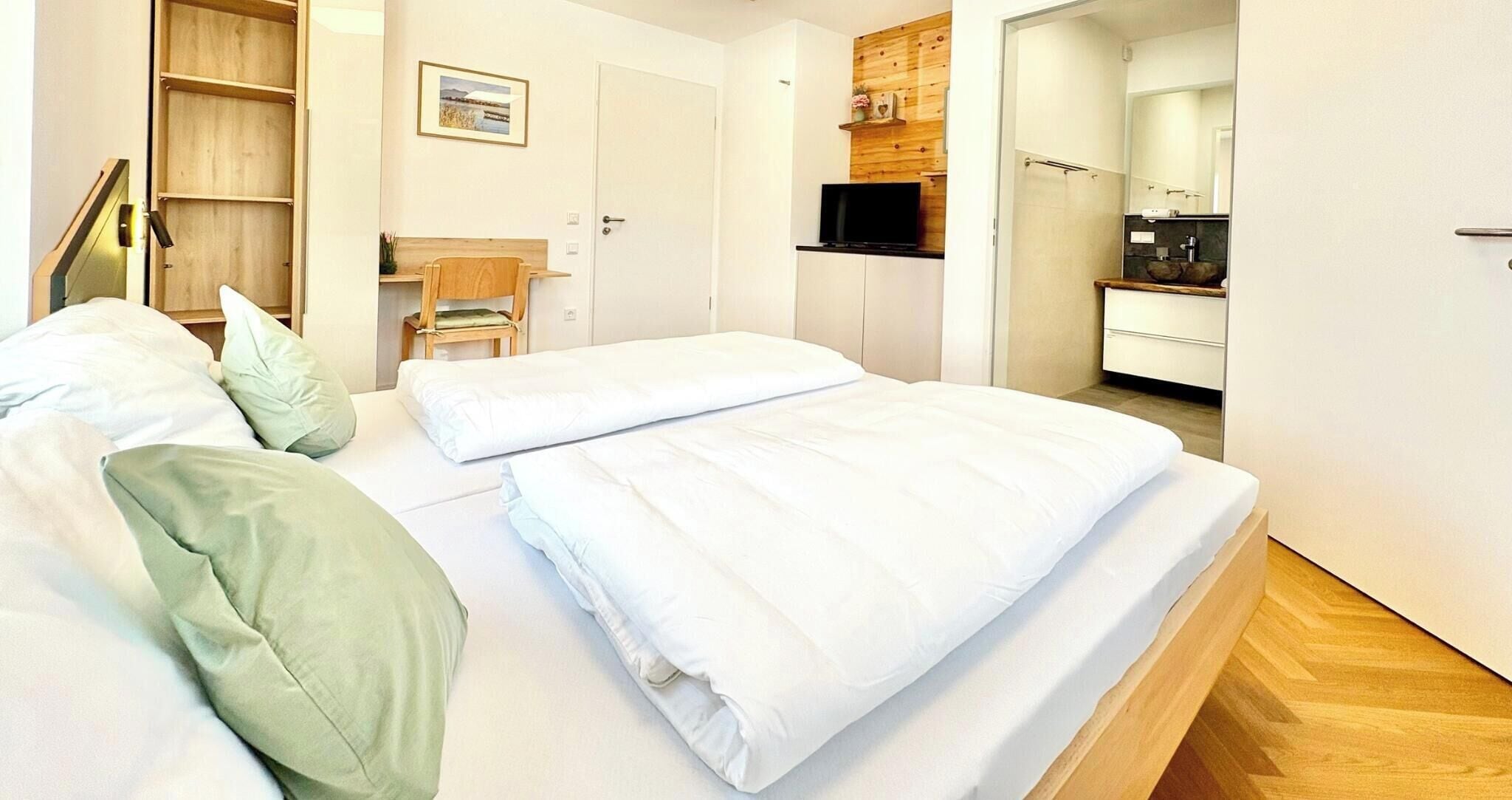 Una cama bien hecha con ropa de cama blanca, una silla de madera y una mesita con una planta.