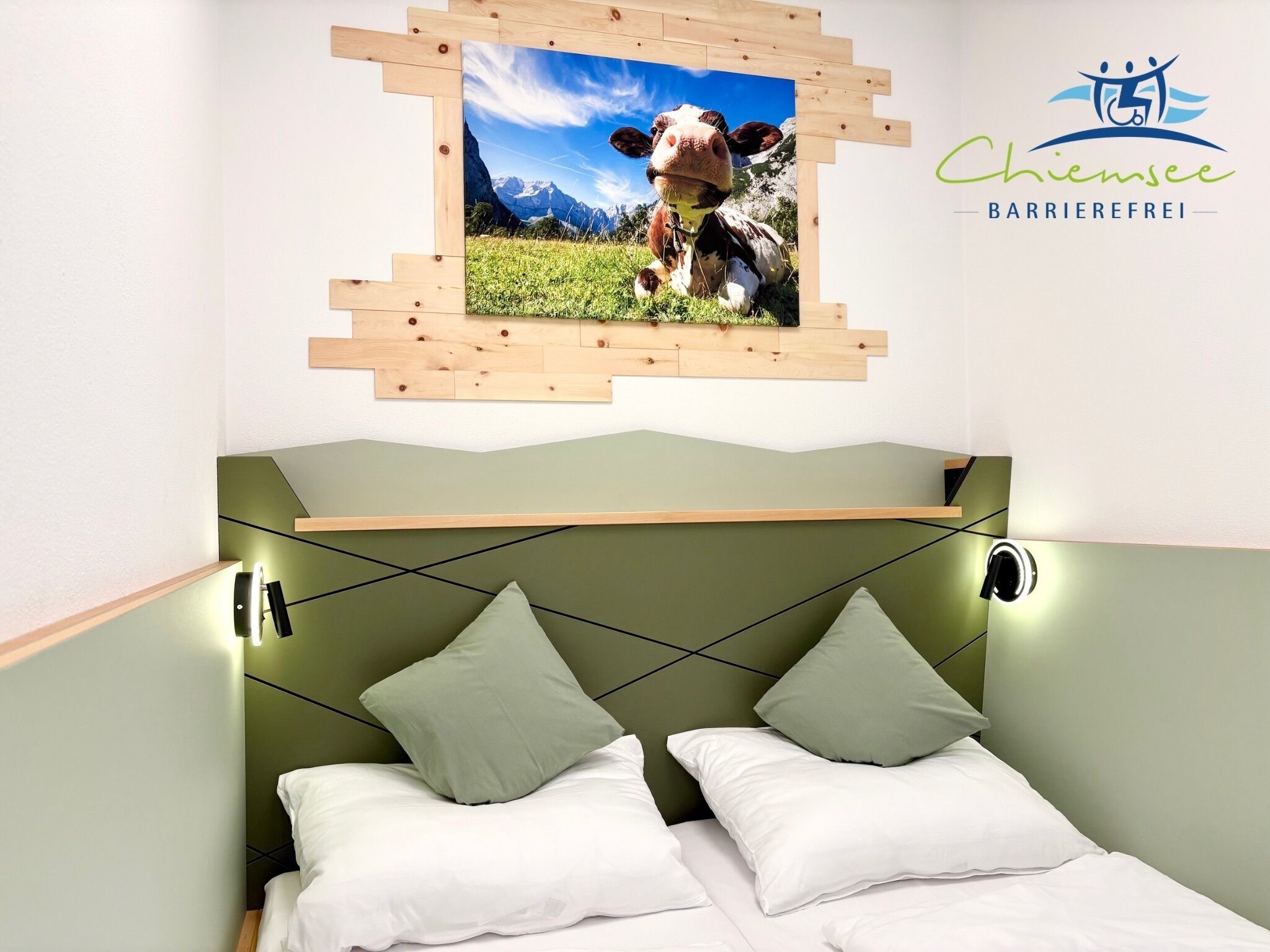 Een slaapkamer met een groen hoofdeinde, twee kussens, een afbeelding van een koe en het logo van 'Chiemsee Barrier-free'.