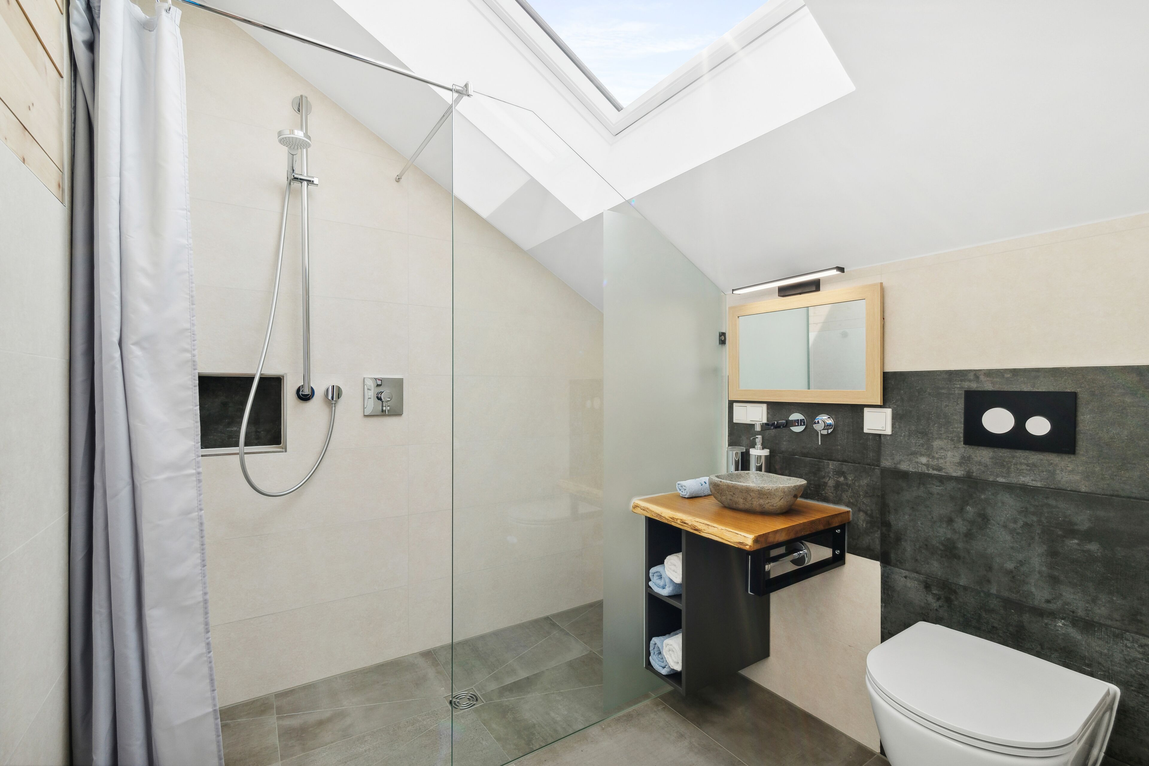 A modern bathroom with a skylight, a glass shower enclosure, a wooden vanity, and a white toilet.