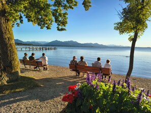Un grupo de personas sentadas en bancos junto a un lago, con montañas al fondo.