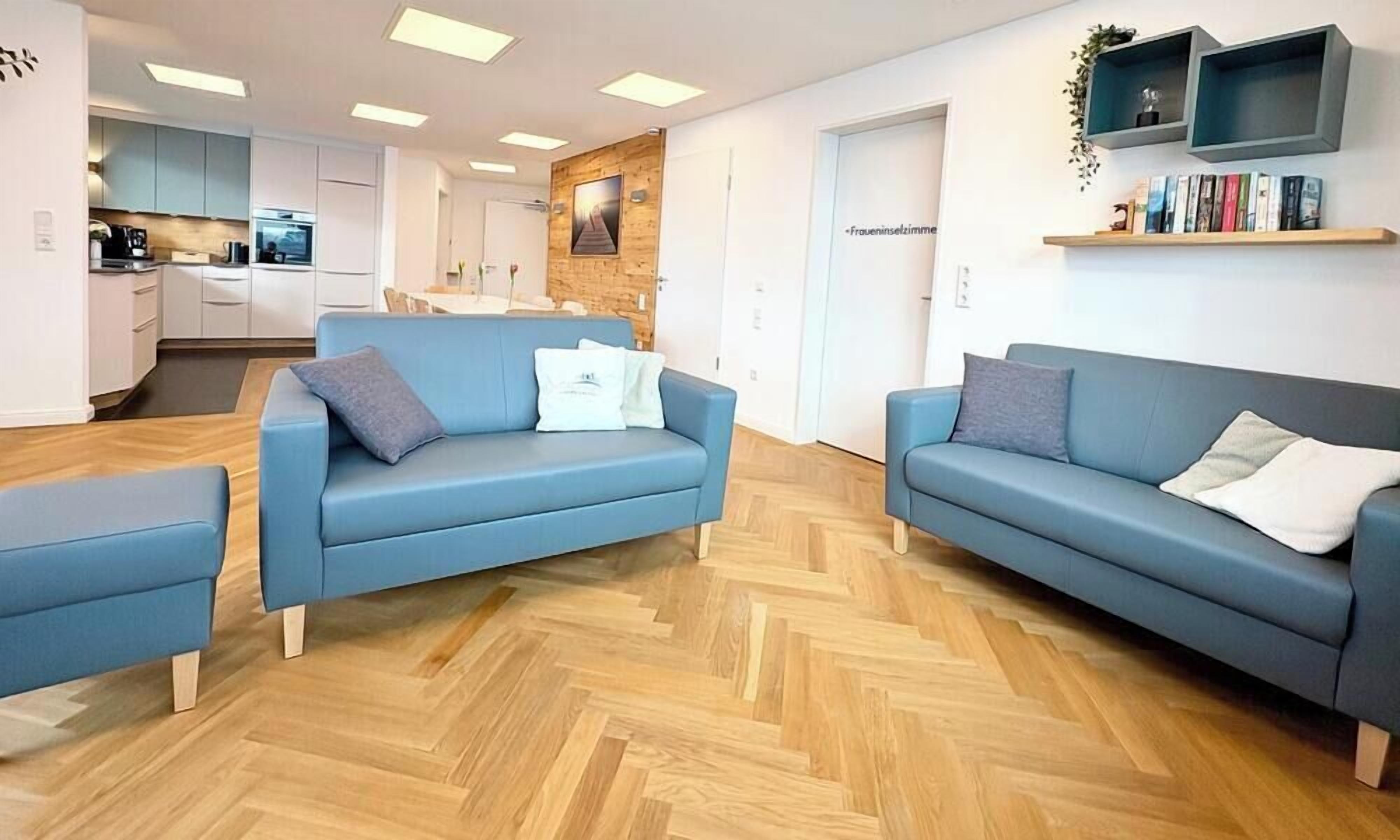 A modern living room with two blue sofas, a wooden herringbone floor, and a kitchen in the background.