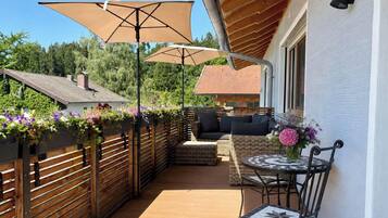 A covered outdoor patio with a wooden fence, a wicker sofa, and a table with a vase of flowers.