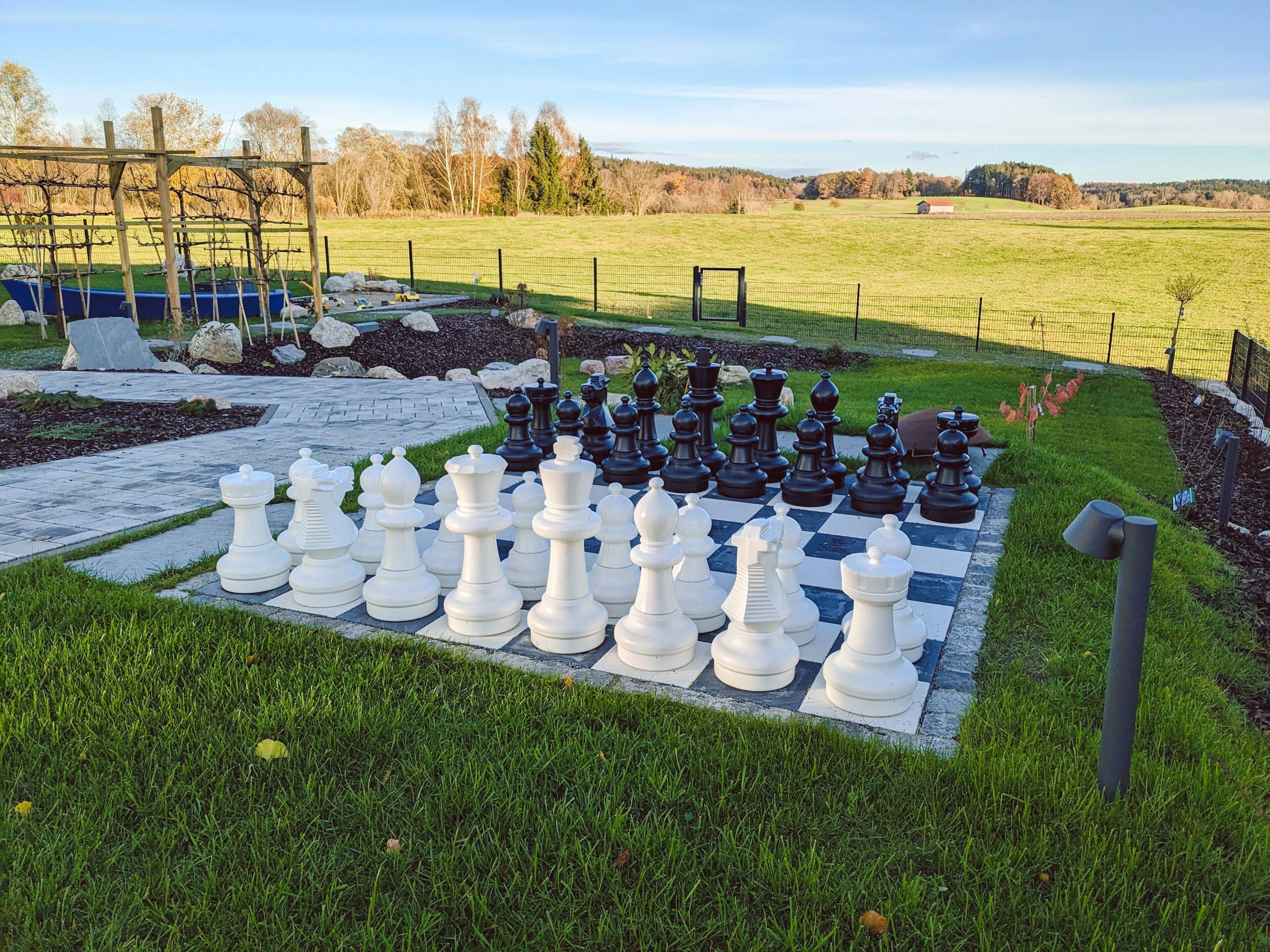 A large outdoor chess set on a paved area surrounded by grass and a fence.