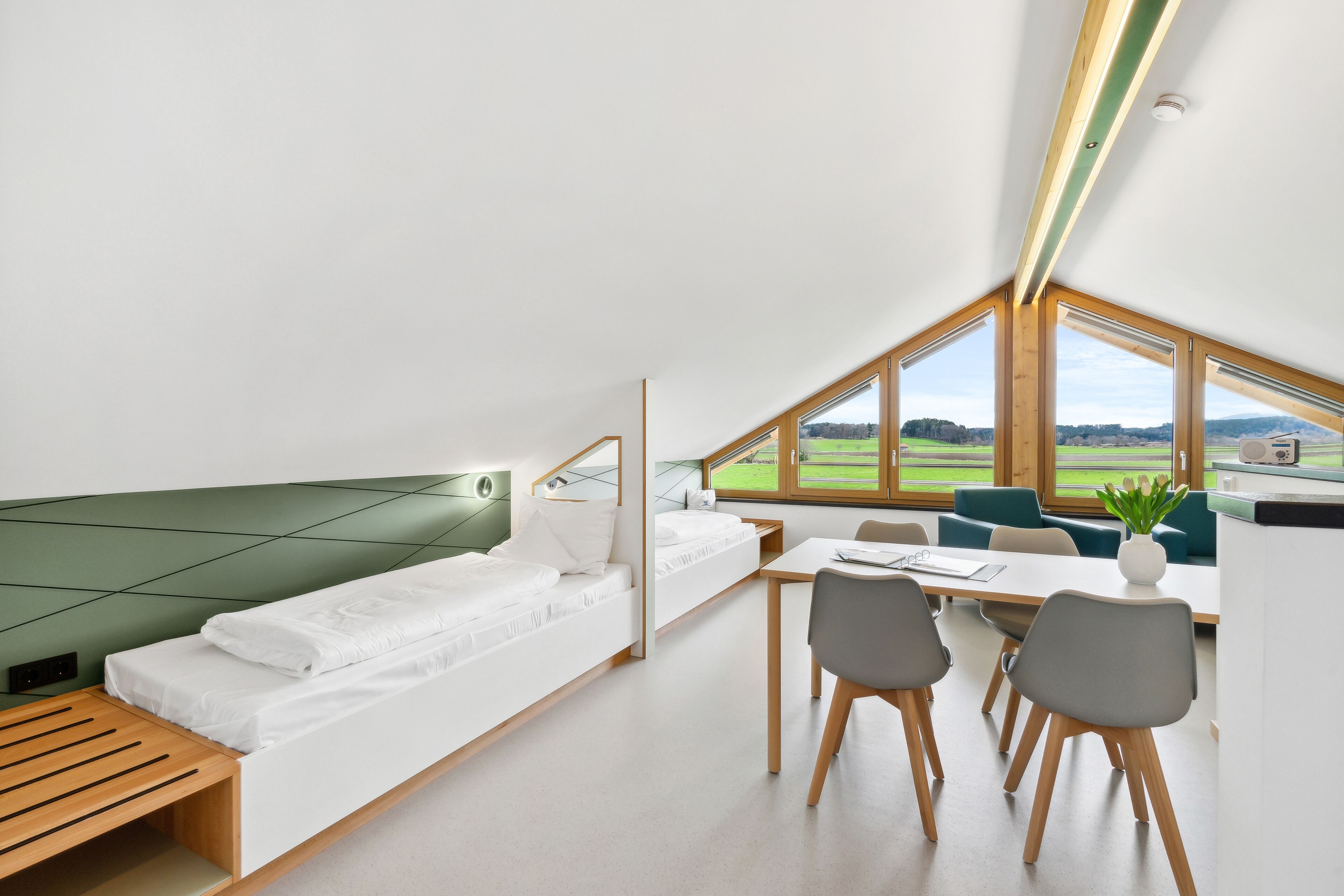 Una habitación moderna y compacta con cama tipo altillo, zona de comedor con mesa y sillas, y grandes ventanales con vistas al campo.
