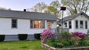 A white two-story house with a front porch, a mailbox, and a well-maintained garden with pink flowers.