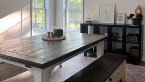 A room with a large table, a ceiling fan, two windows, and a black shelving unit with various items.