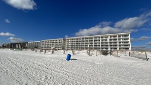 Un hôtel en bord de mer, avec plusieurs étages et des balcons, une plage de sable en premier plan et un ciel bleu dégagé.