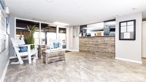 Un hall d’entrée spacieux avec un comptoir de réception en bois, un coin salon avec un fauteuil blanc et une petite table, et un sol carrelé.