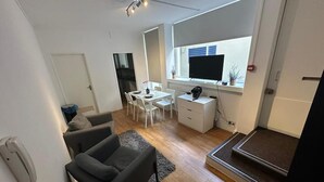 A modern living room with a grey sofa, a dining area with a white table and chairs, a small white desk with a computer, and a large window with blinds. - Charming Studio in Bond Street (London)
