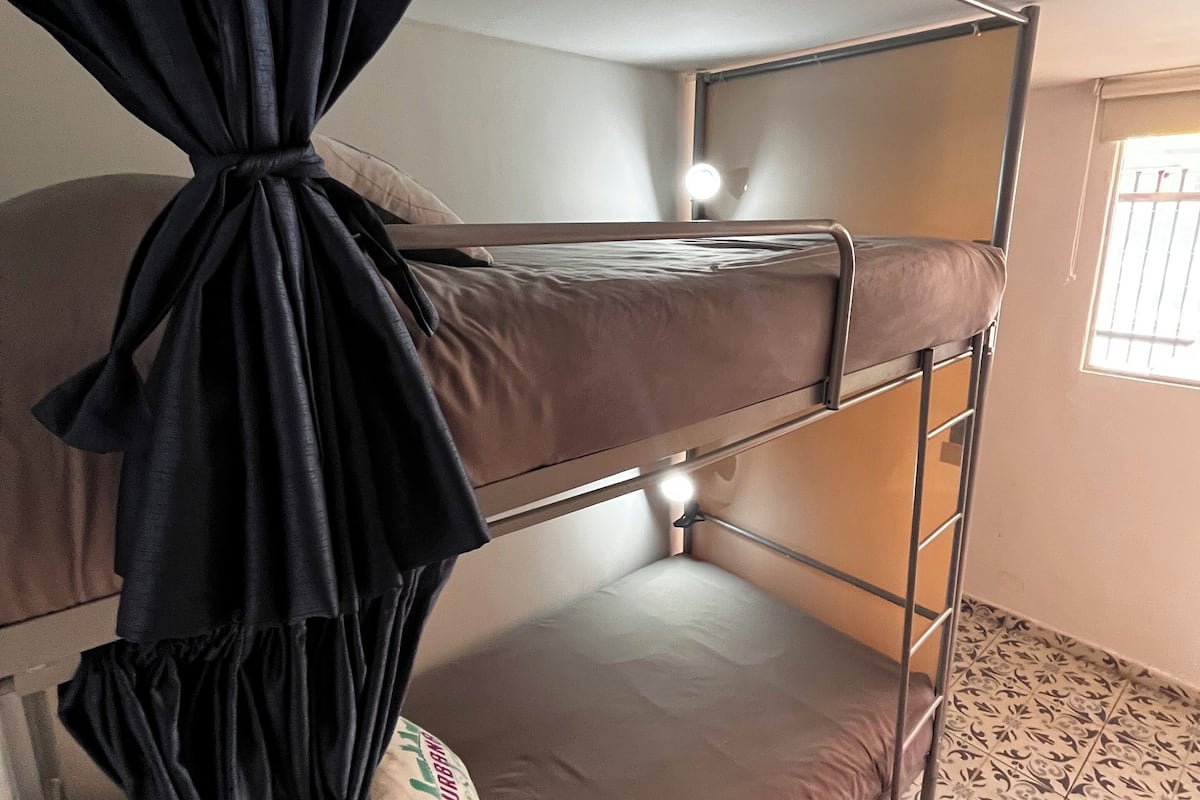 Cama litera con una cortina atada en la parte superior, una cama individual abajo y una ventana con barrotes.