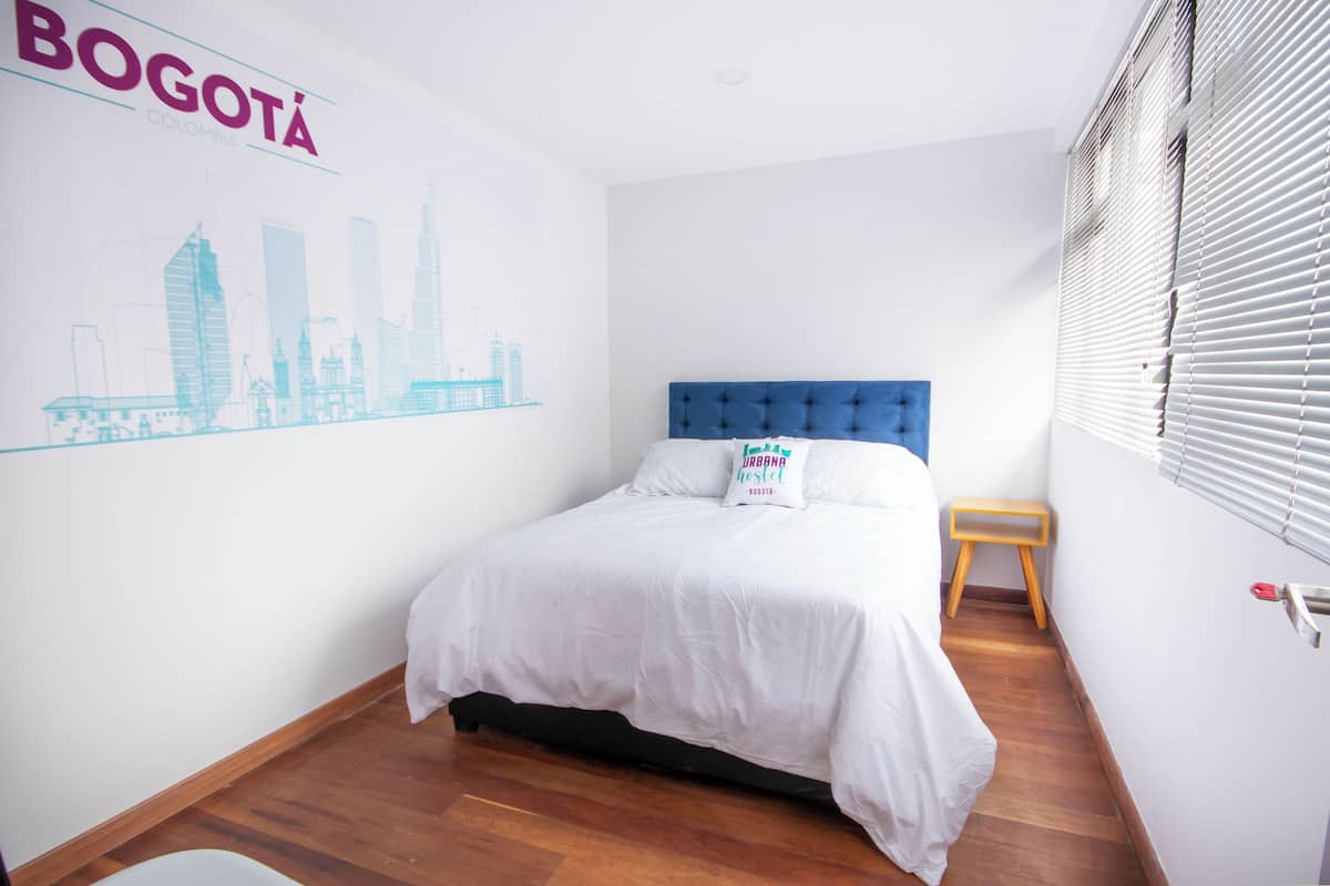 Un dormitorio con una cama, un taburete de madera, una pared con una ilustración de un paisaje urbano y una ventana con persianas.