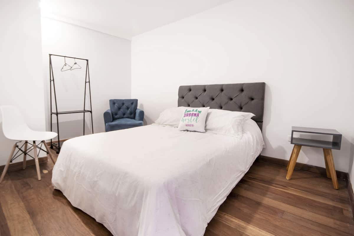 Un dormitorio ordenado con una cama grande, una silla, una mesita y un perchero.