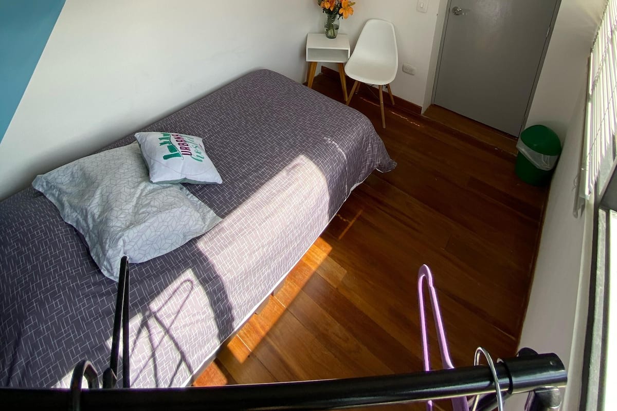 Una habitación pequeña y bien iluminada con una cama, un escritorio, una silla y una puerta.