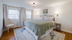 Ein Schlafzimmer mit einem Bett, einem Sessel, einem Nachttisch, einer Lampe, einer Deckenleuchte und einem Fenster mit Vorhängen.