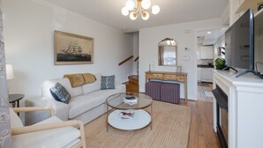 Ein modernes Wohnzimmer mit einem weißen Sofa, einem Glastisch und einem Essbereich. Im Hintergrund ist eine Küche zu sehen.