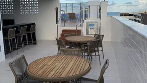Una terraza en la azotea con una mesa de madera redonda y sillas de mimbre, una zona de bar con taburetes y vistas al mar.