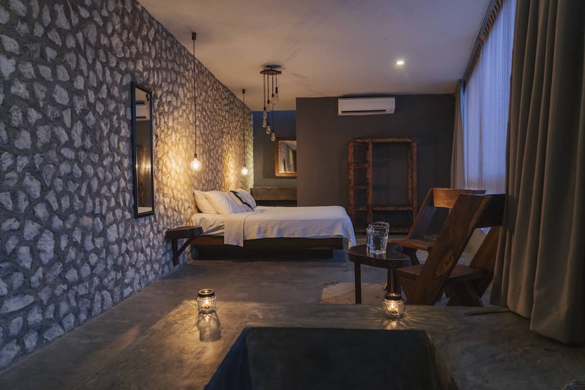 Un dormitorio con pared de piedra, una cama, una silla de madera, una mesita y una lámpara colgante.