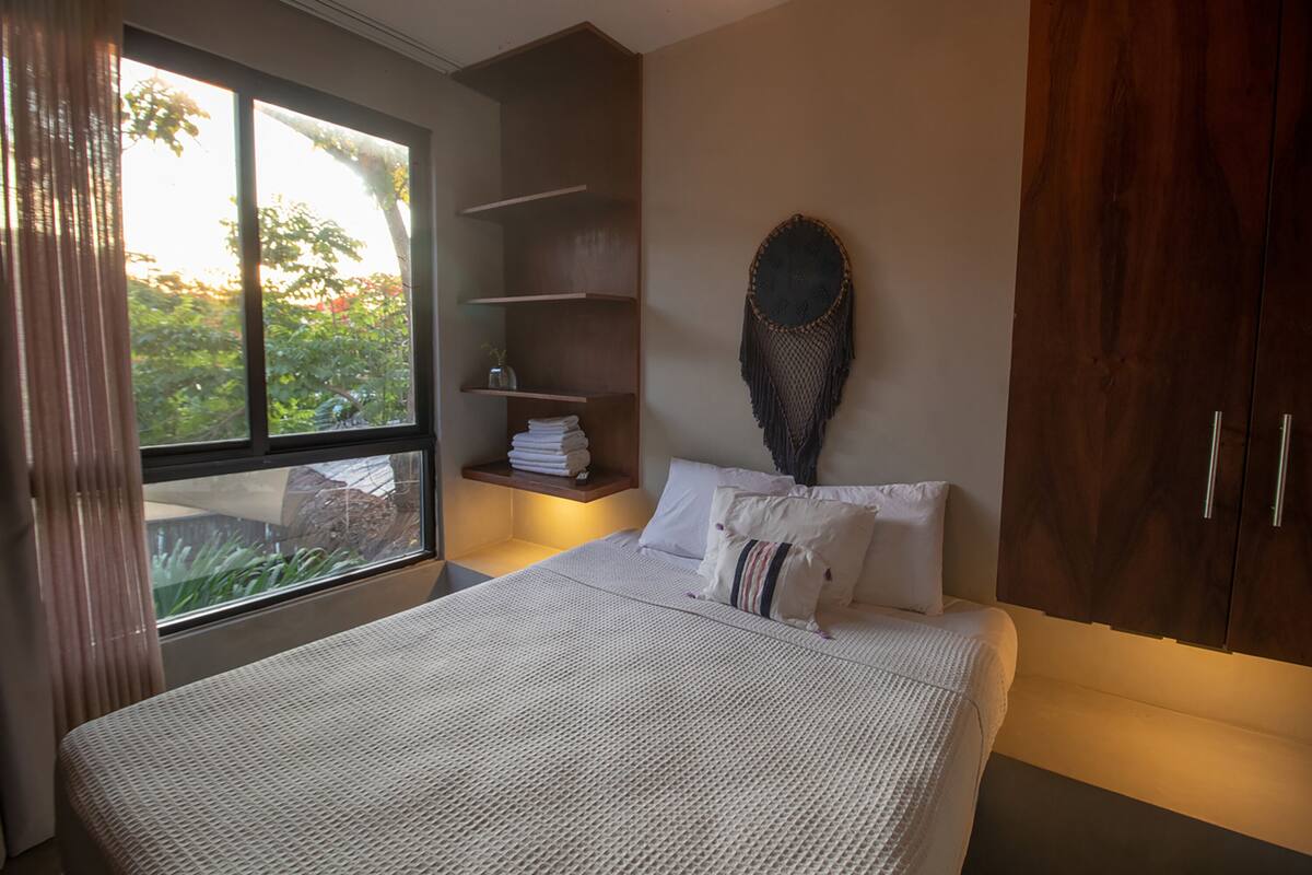 Un dormitorio con un ventanal grande, una cama con ropa de cama blanca, un armario de madera y un estante con toallas dobladas.