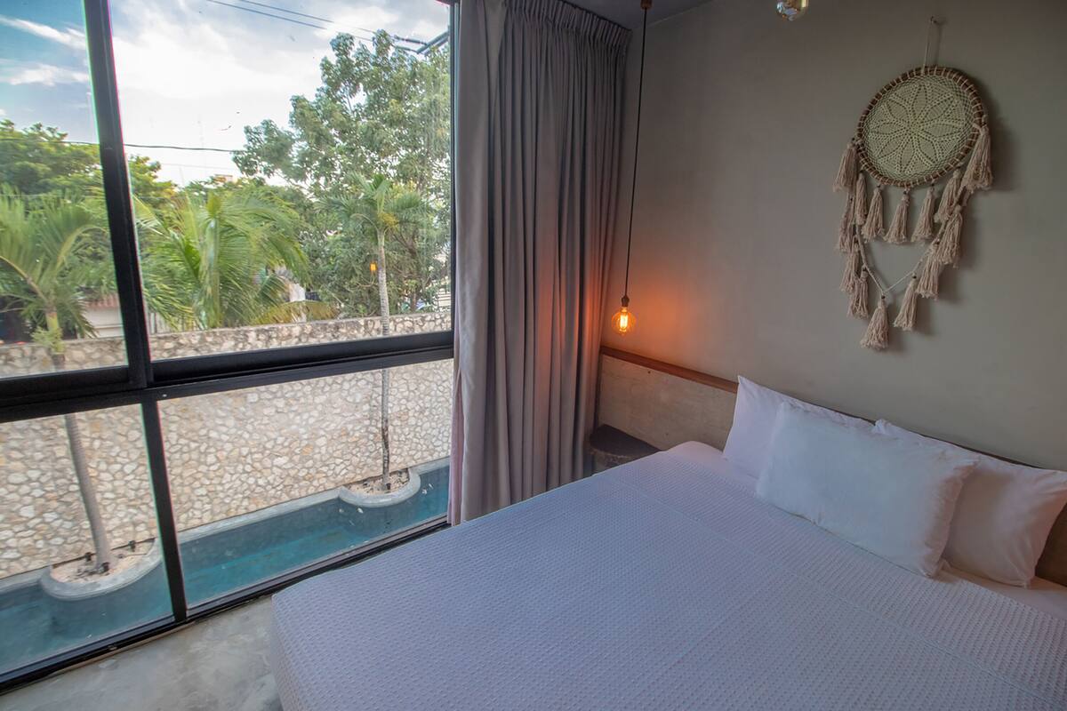 Un dormitorio con una cama grande, una ventana con vista a follaje y un atrapasueños en la pared.