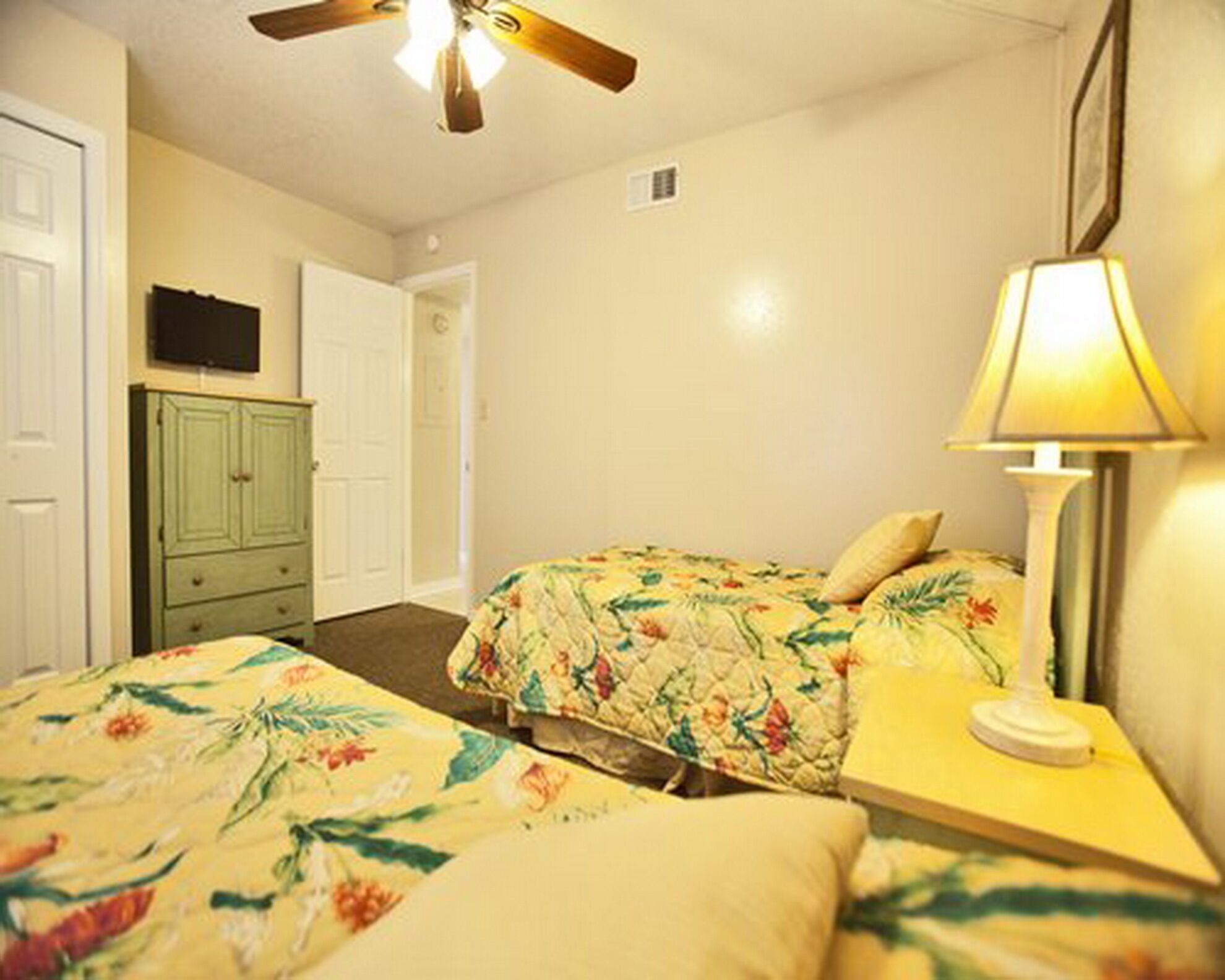 Une chambre avec deux lits, un ventilateur de plafond, une table de chevet avec une lampe, une commode verte et une porte.