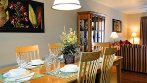 Un comedor con una mesa de madera dispuesta para cuatro personas, rodeada de sillas. Hay una planta en maceta sobre la mesa, y un cuadro grande encima. Se observa un mueble con puertas de vidrio al fondo.