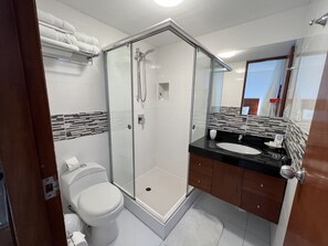 Un baño moderno con ducha envuelta en vidrio, inodoro blanco y lavamanos negro con espejo superior.