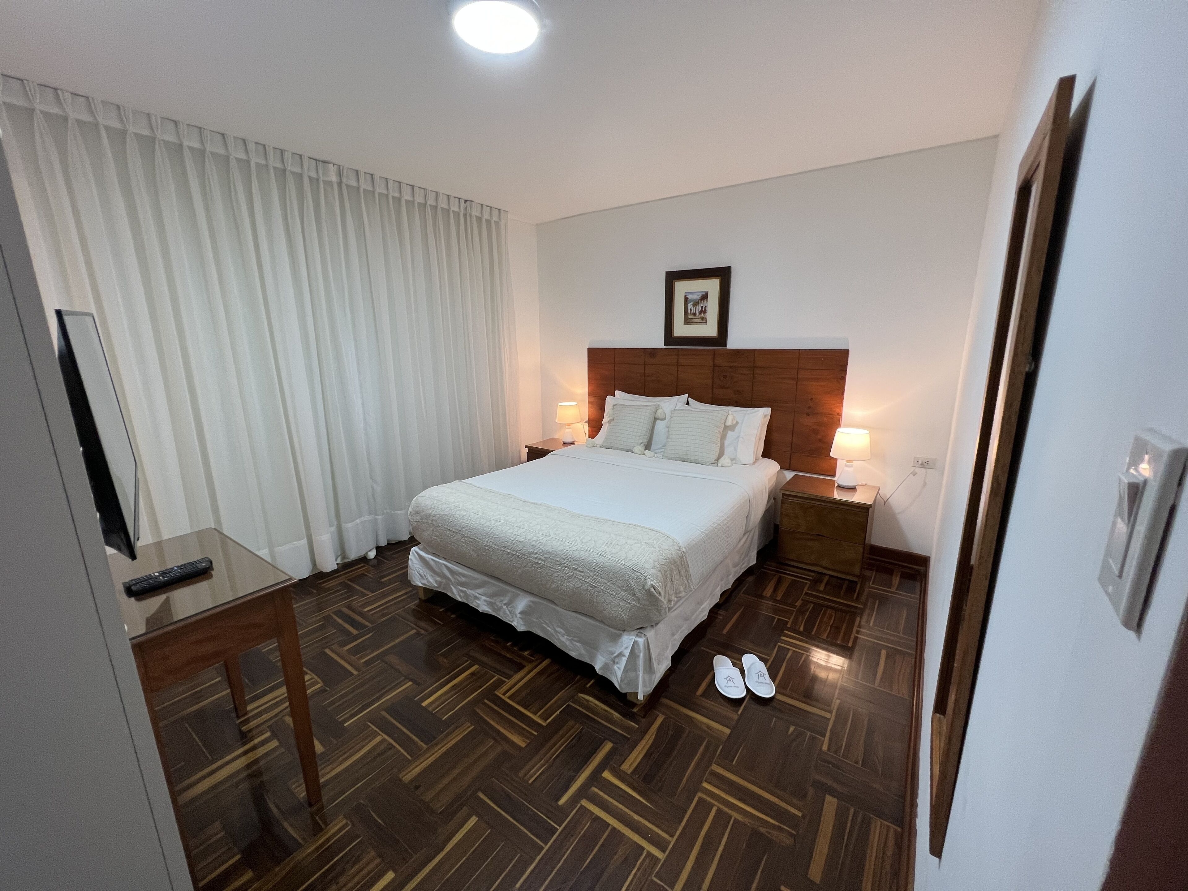 A hotel room with a large bed, a bedside table, a TV, and a pair of slippers on the floor.