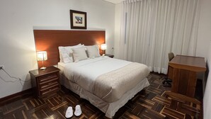 Ein Hotelzimmer mit Holz-Kopfteil, einem Bett mit weißen Laken, zwei Nachttischlampen, einem Schreibtisch und einem Sessel.