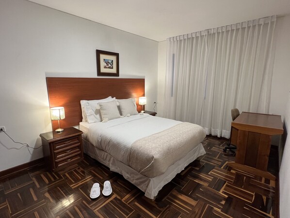 Habitación de hotel con cabecera de madera, cama con ropa blanca, dos lámparas de noche, un escritorio y una silla.