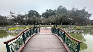 Eine Holzbrücke mit aufwendigen Metallgeländern, die zu einem von Bäumen umgebenen Teich führt.