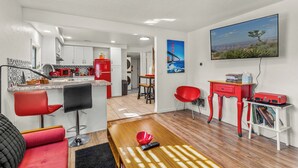 A modern kitchen with white cabinets, a red microwave, and a red refrigerator. There is a dining area with red chairs and a wooden table. A painting of a bridge is on the wall, and a television is mounted above it. - Route 66 Downtown Burgundy Bungalow w/Fenced Yard, AC, W/D, 3 Smart TVs, Wi-Fi (Williams)